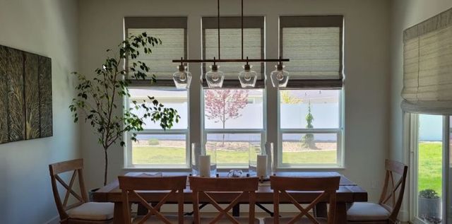 Dining room with wood table, chairs, and large windows with shades, overlooking a backyard.