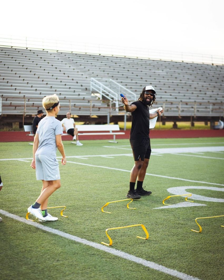 Child sprinting on a sports field, wearing a black shirt and red shorts, with a cone in the foreground.