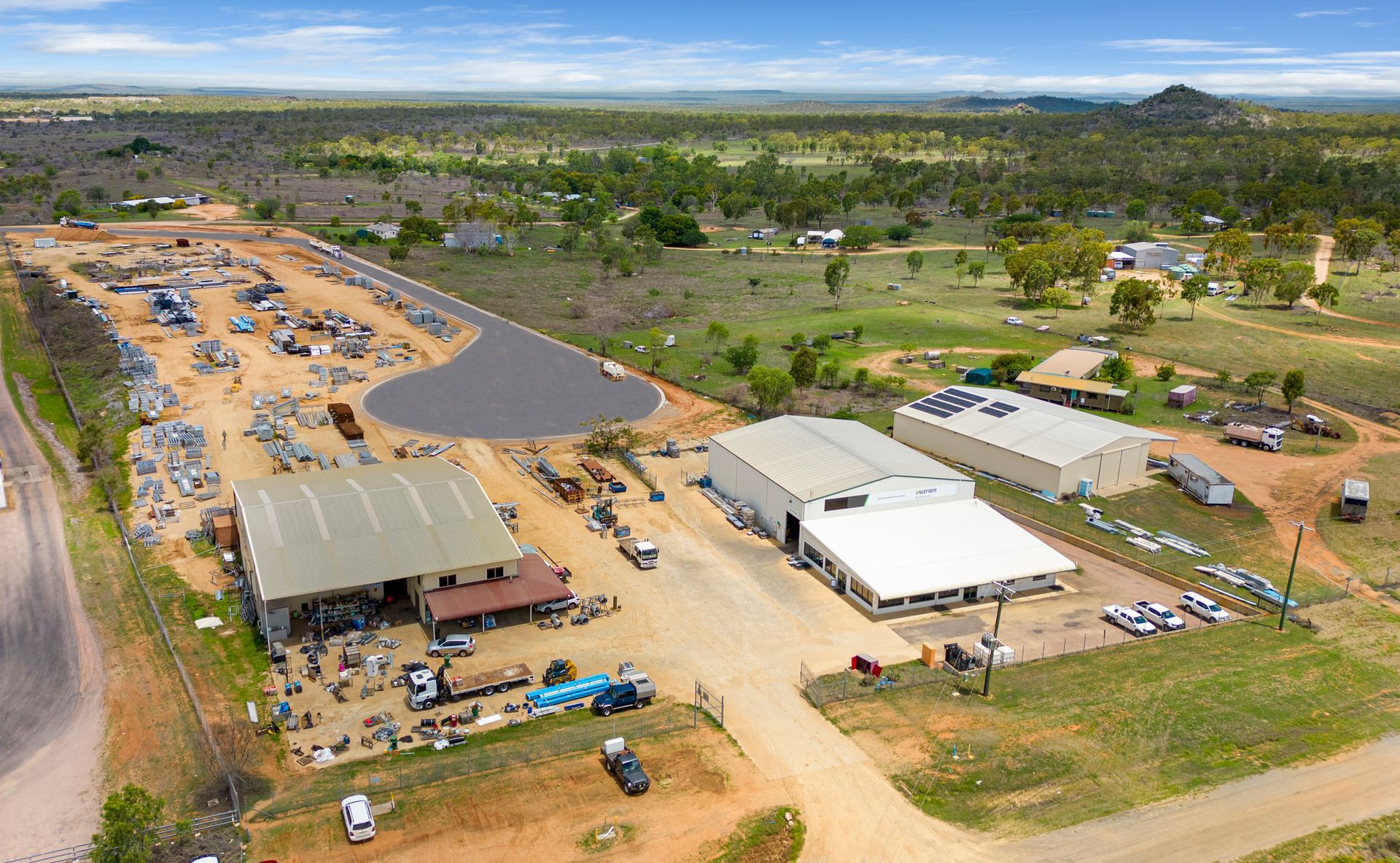Aerial view of a metal recycling facility with buildings, a gravel area, and scattered materials, set against a rural landscape.