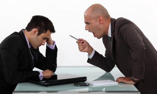 Um homem está gritando com outro homem que está sentado em uma mesa com um laptop.