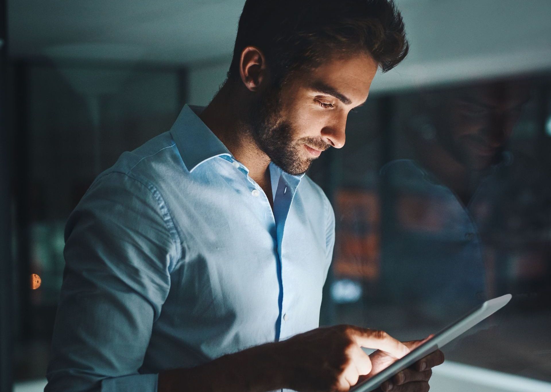 Um homem de camisa azul está usando um tablet à noite.