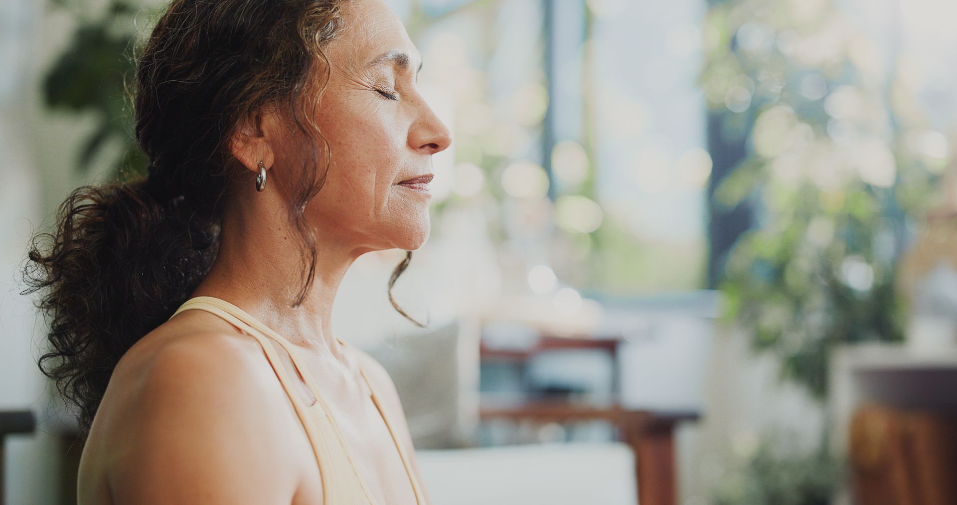 Woman in profile meditating indoors near a bright window, eyes closed in a calm pose.