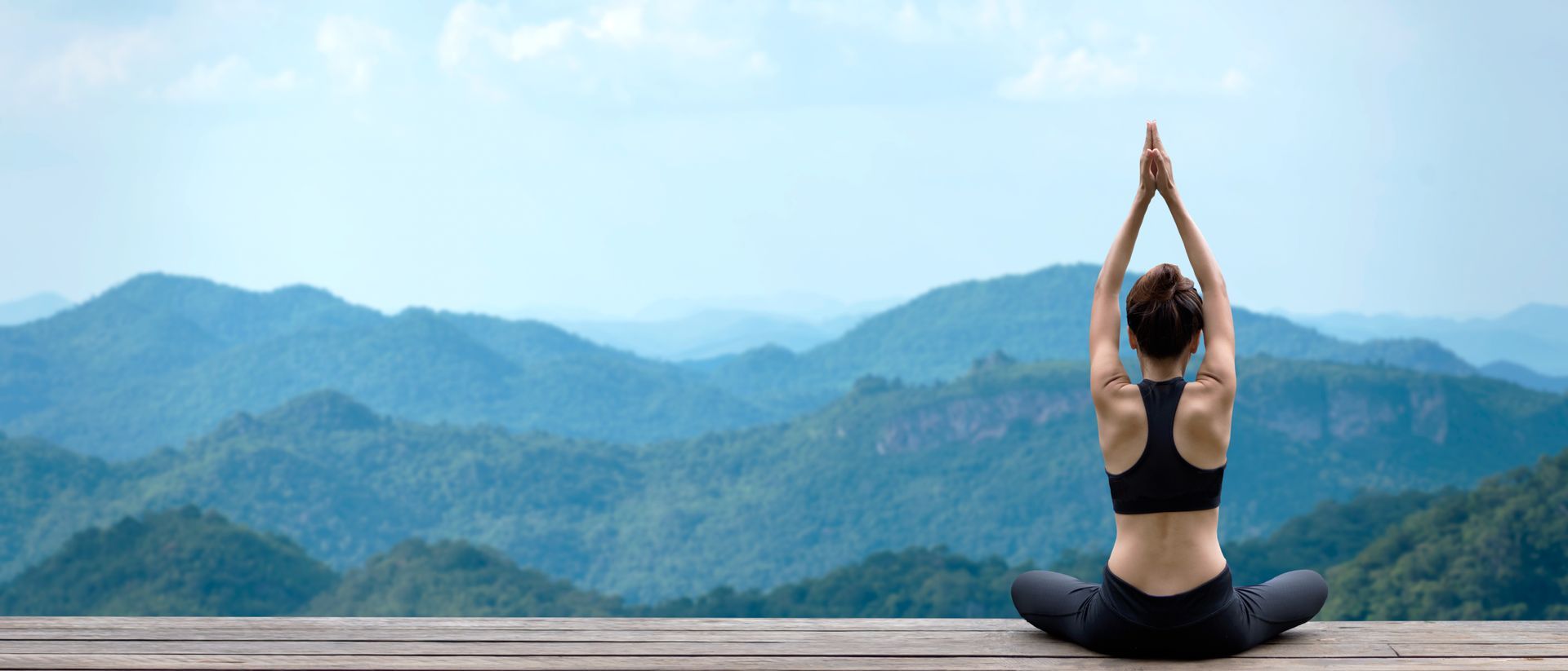 Person seated in lotus pose with arms raised, overlooking misty blue mountain ridges at sunrise
