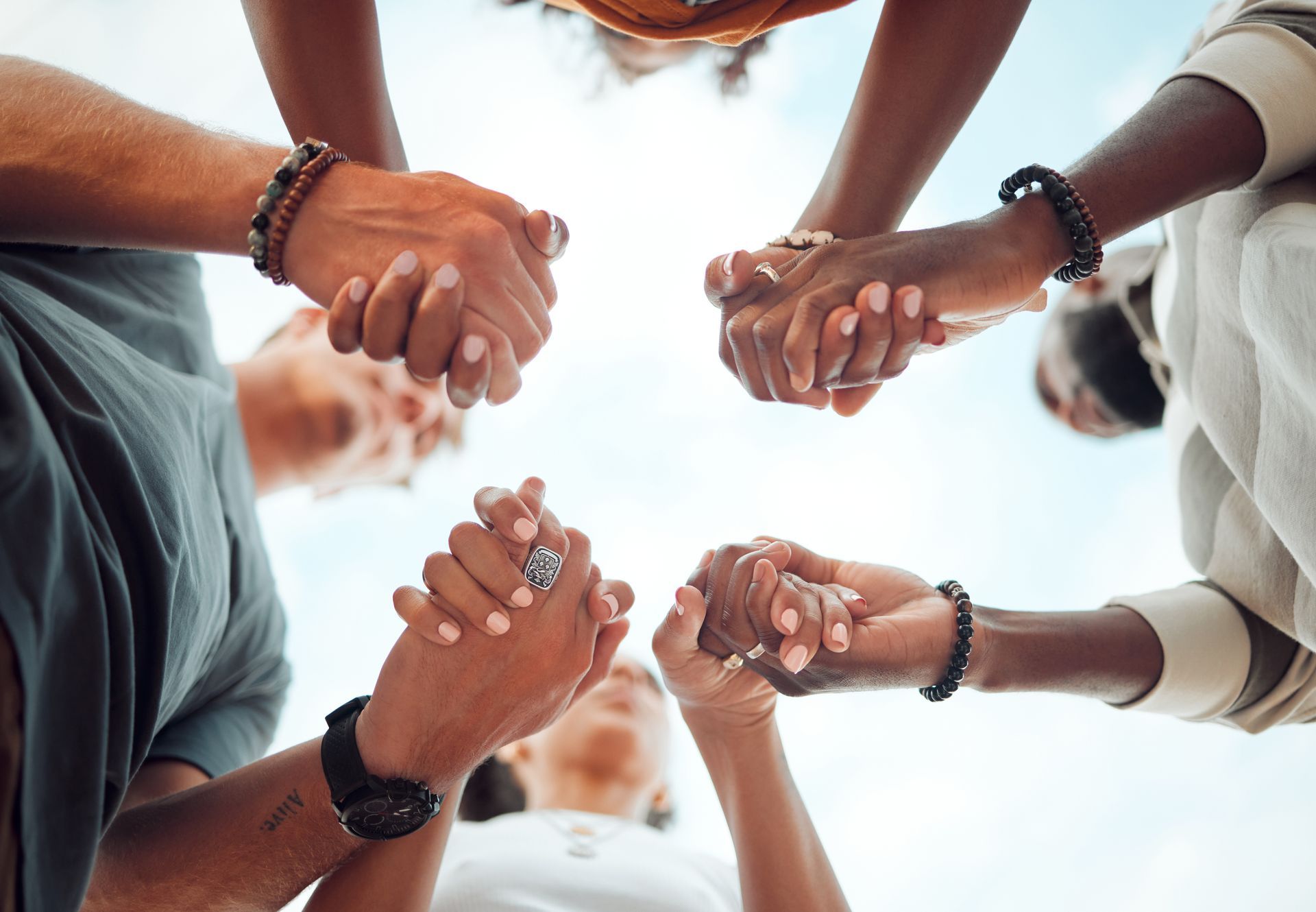 Several people join hands in a circle, wearing bracelets, viewed from below against a bright sky.