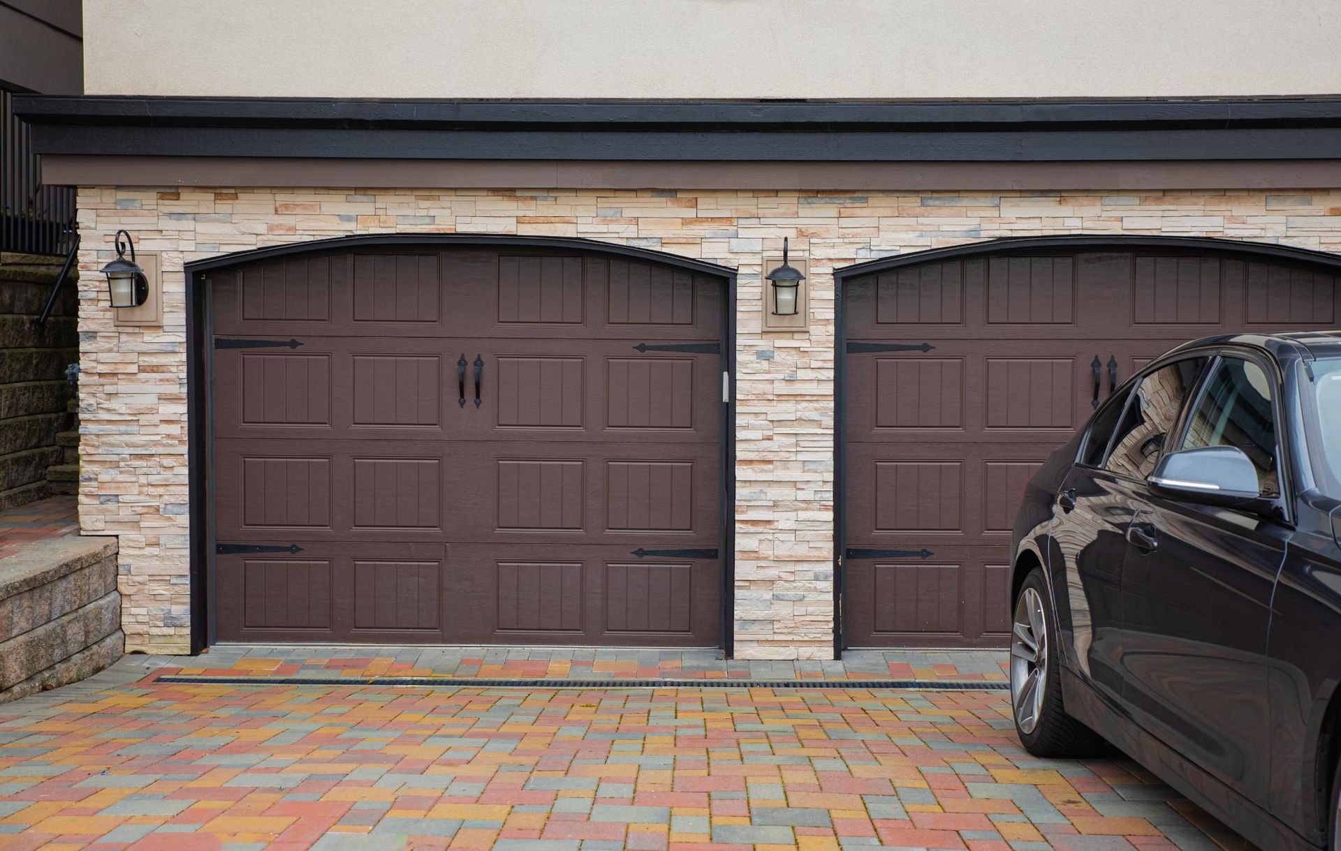 Two brown garage doors on a stone house, with a black car parked in front on a brick driveway.