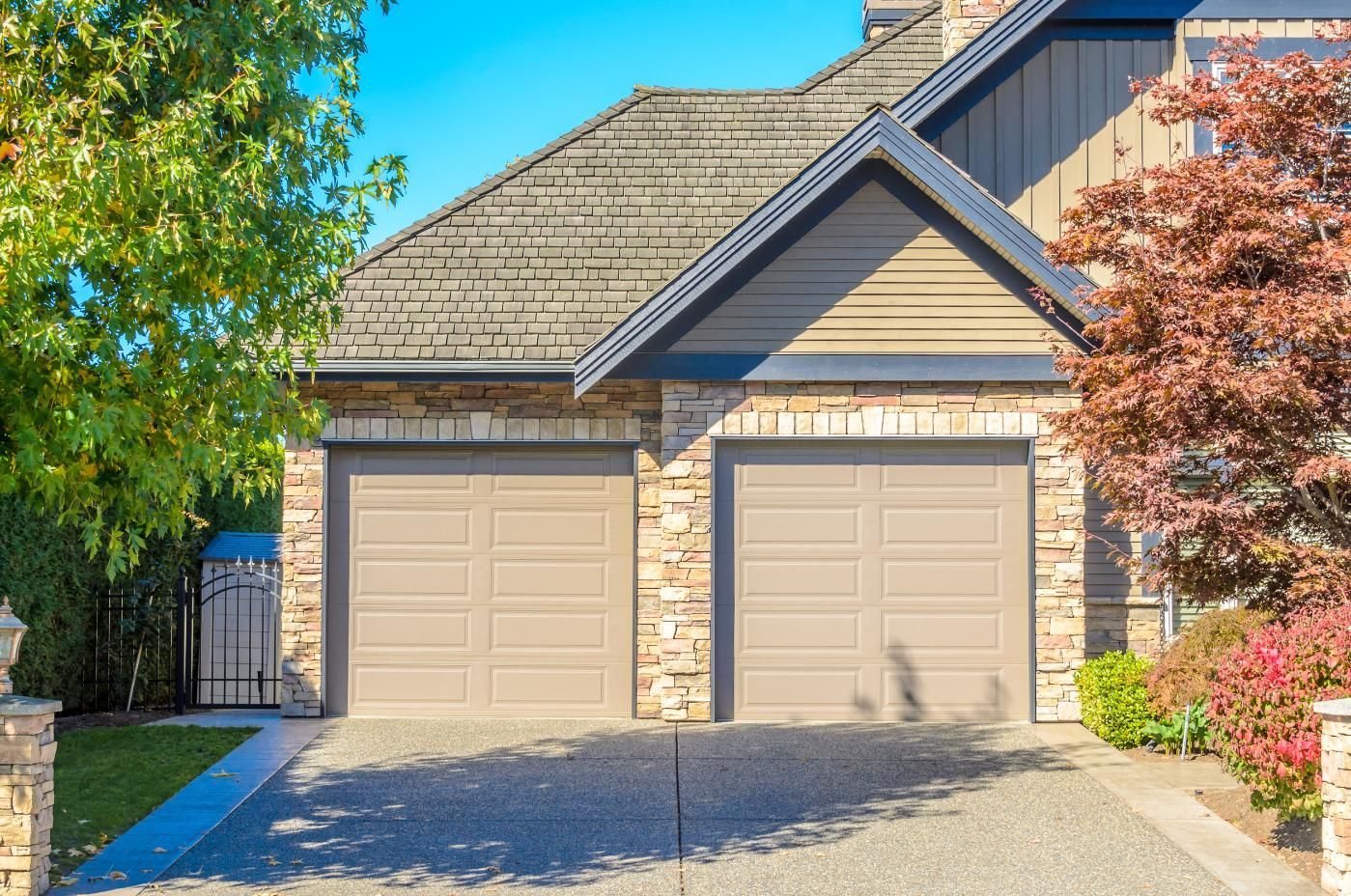 Suburban house with two beige garage doors, gray roof, and trees in the front yard.