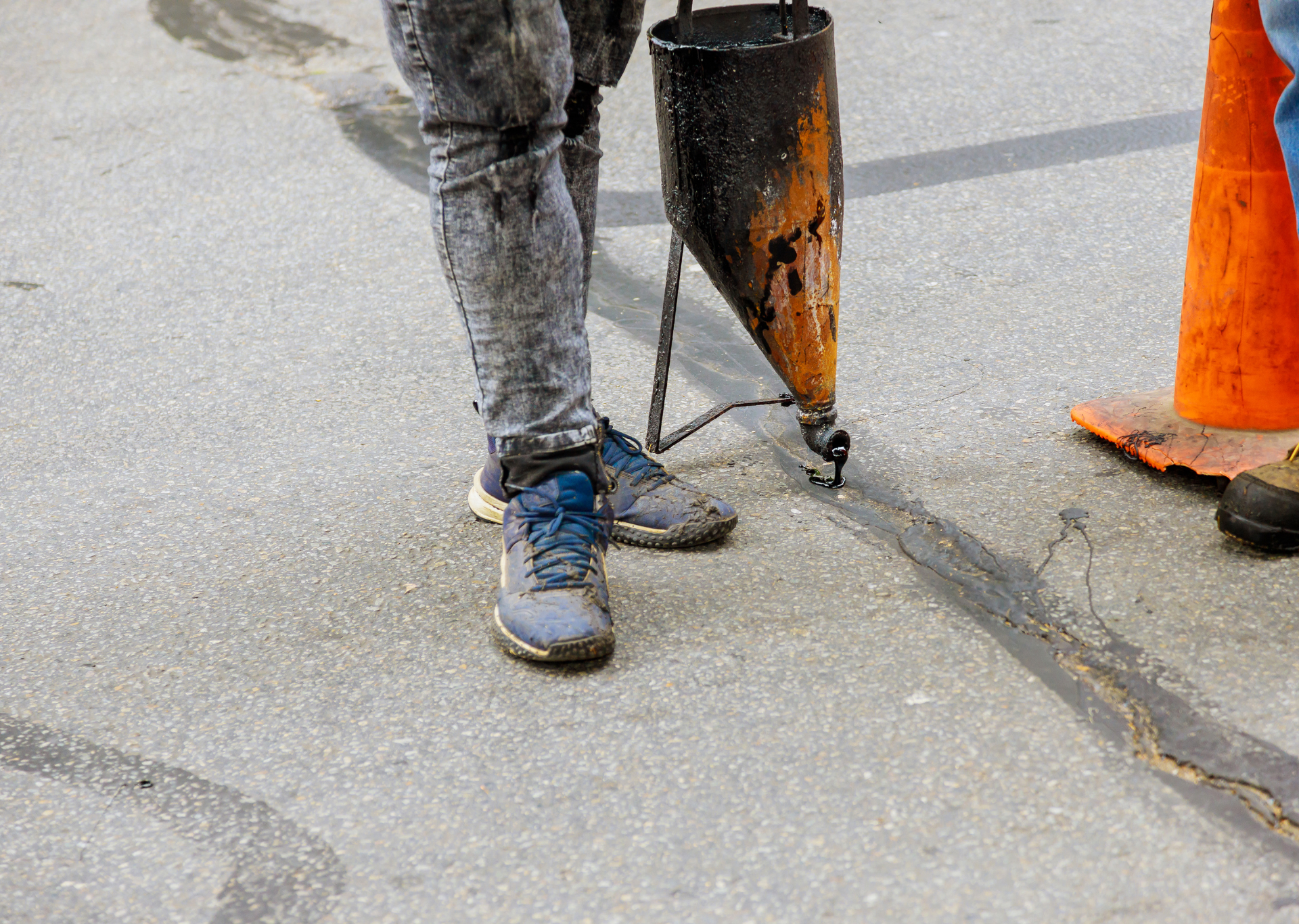 Person in gray jeans and blue sneakers applying sealant to an asphalt crack with a black applicator.