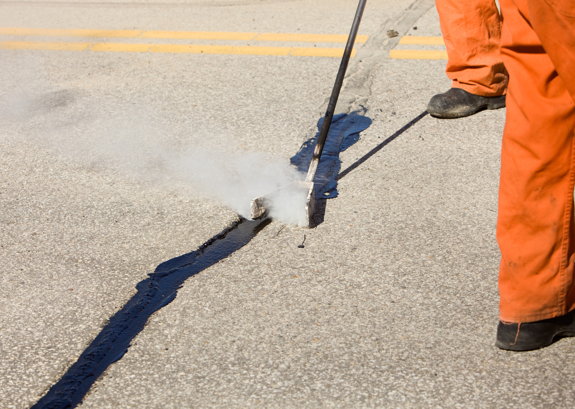 Person in orange jumpsuit seals road crack with hot asphalt.