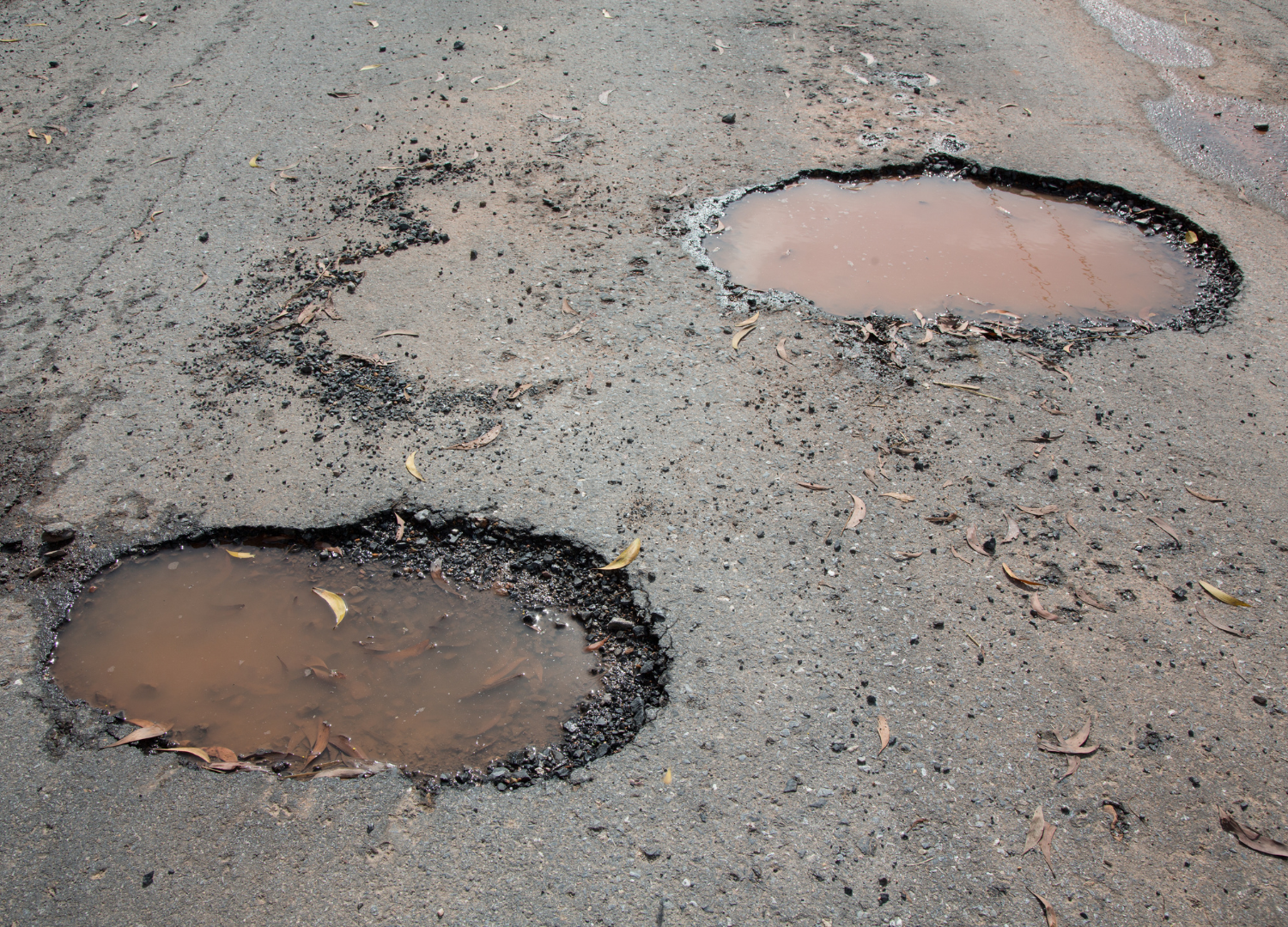 Two water-filled potholes on a gray asphalt road. Dark edges surround the brown water.