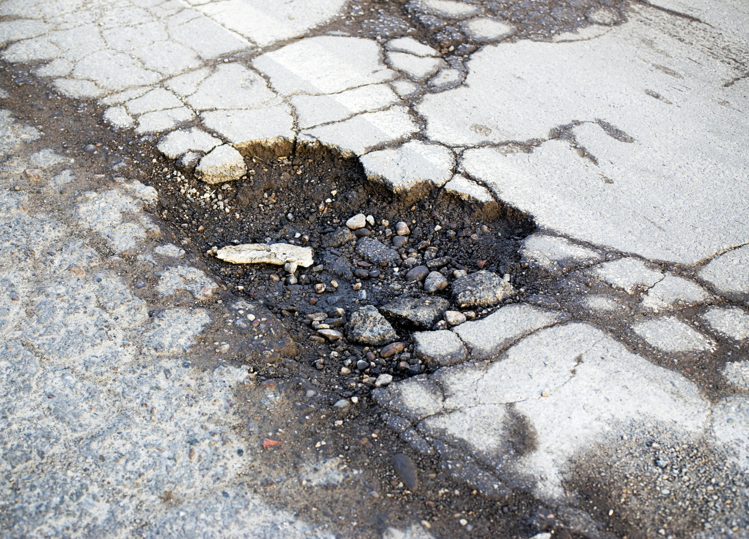 Pothole in cracked asphalt road surface. Dark asphalt and loose rocks are visible within the hole.