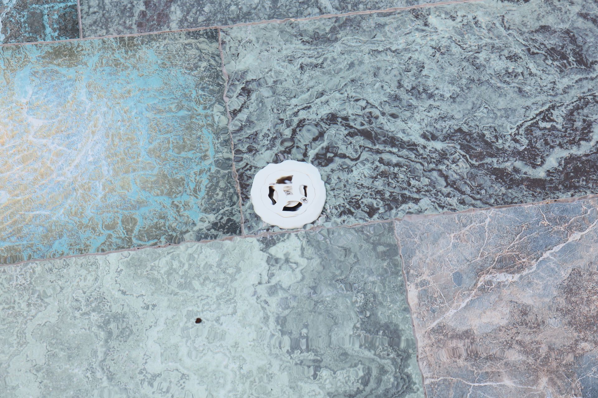 Close-up of patterned stone tiles in shades of blue, green, and gray, with a white drain cover in the center.