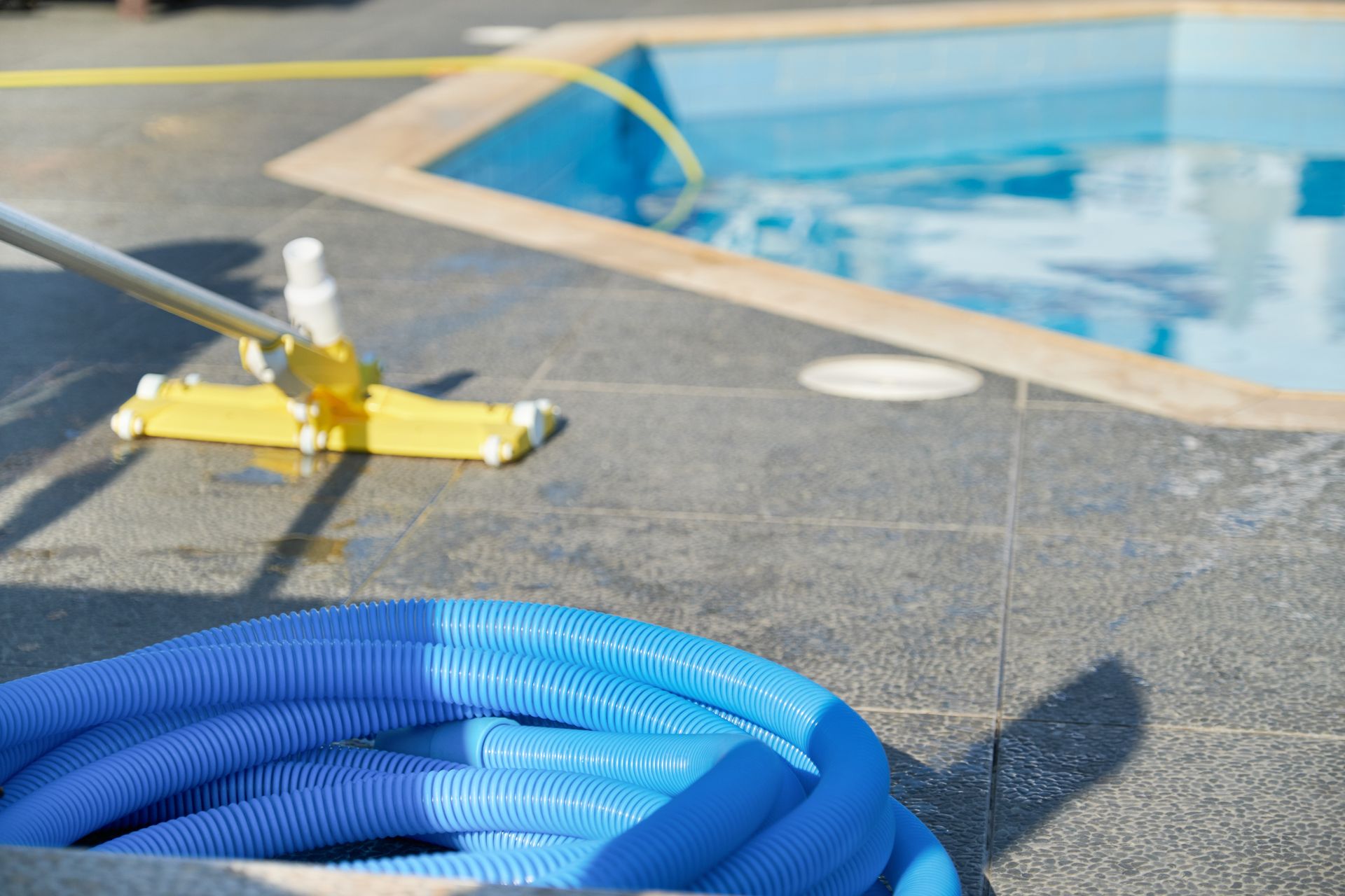 A person cleaning a blue pool with a yellow brush and blue hose on a gray patio.
