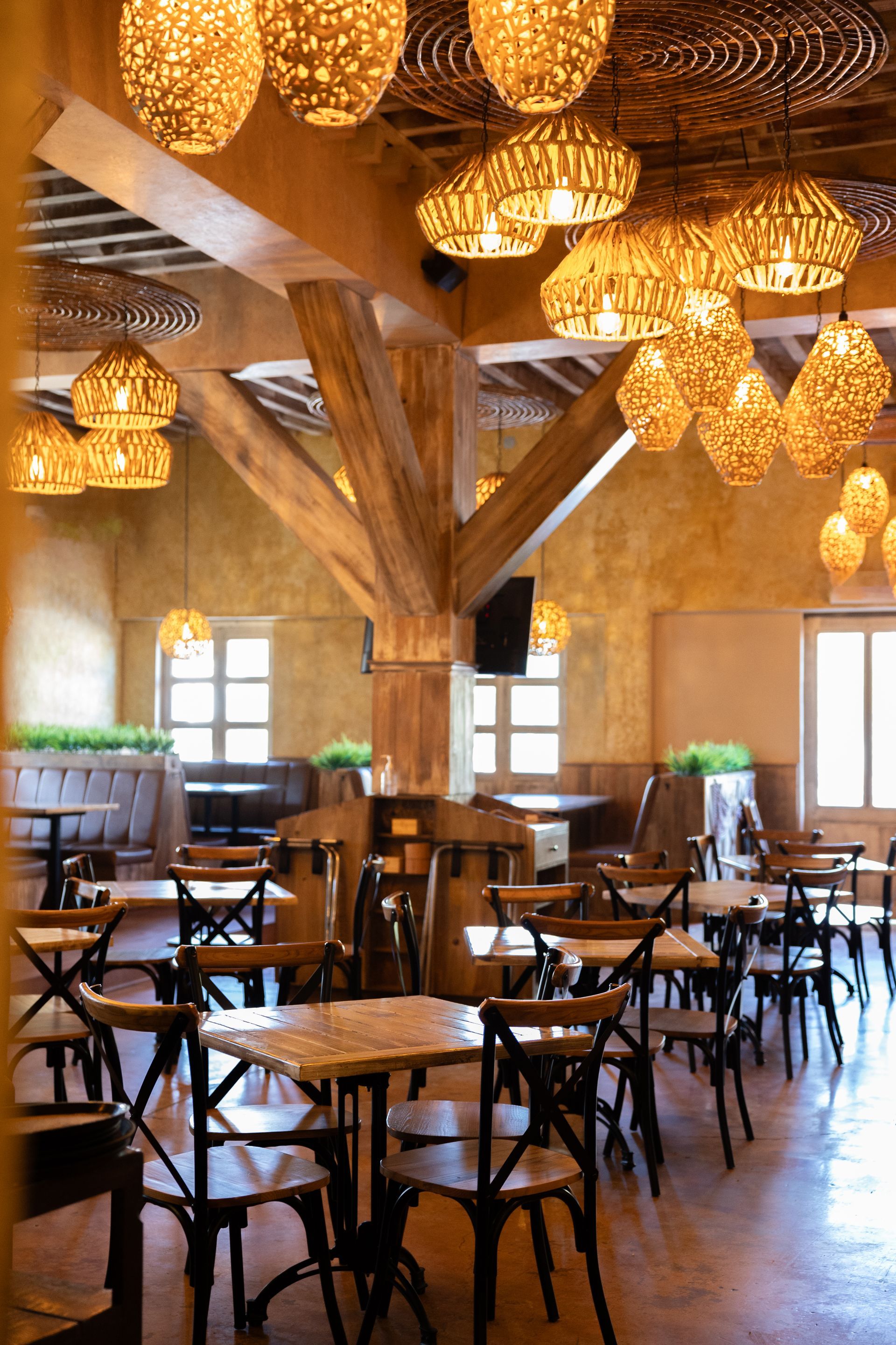 Interior de restaurante con mesas y sillas de madera, lámparas tejidas decorativas y una gran viga de soporte de madera.