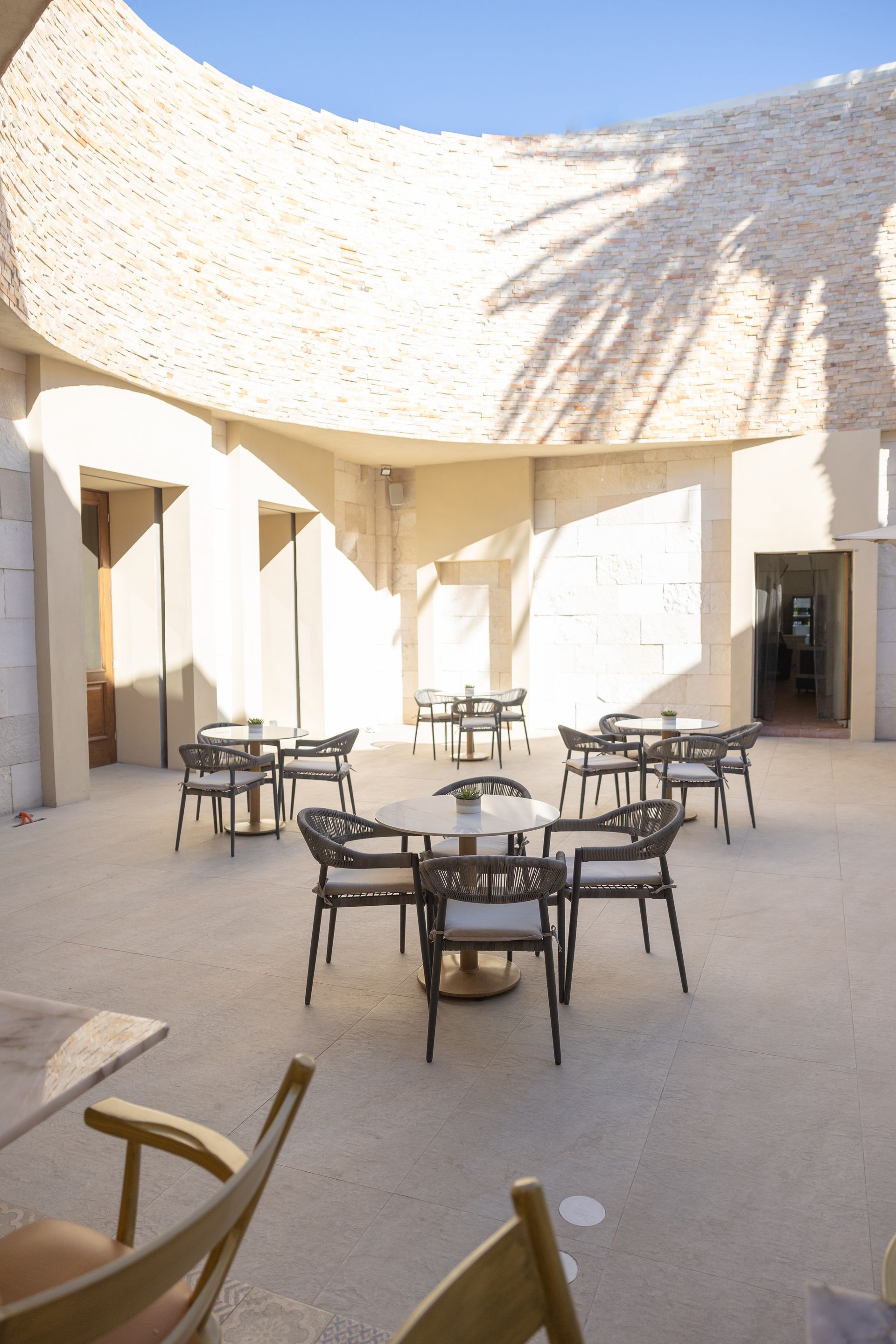 Comedor al aire libre con mesas y sillas en un patio de color arena, bajo un techo curvo con textura.