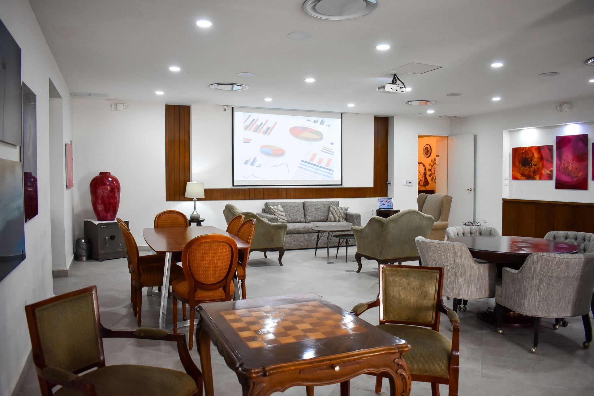 Una sala de estar bien iluminada con sofás, sillas, mesas y un tablero de ajedrez. Una pantalla de proyección muestra imágenes.