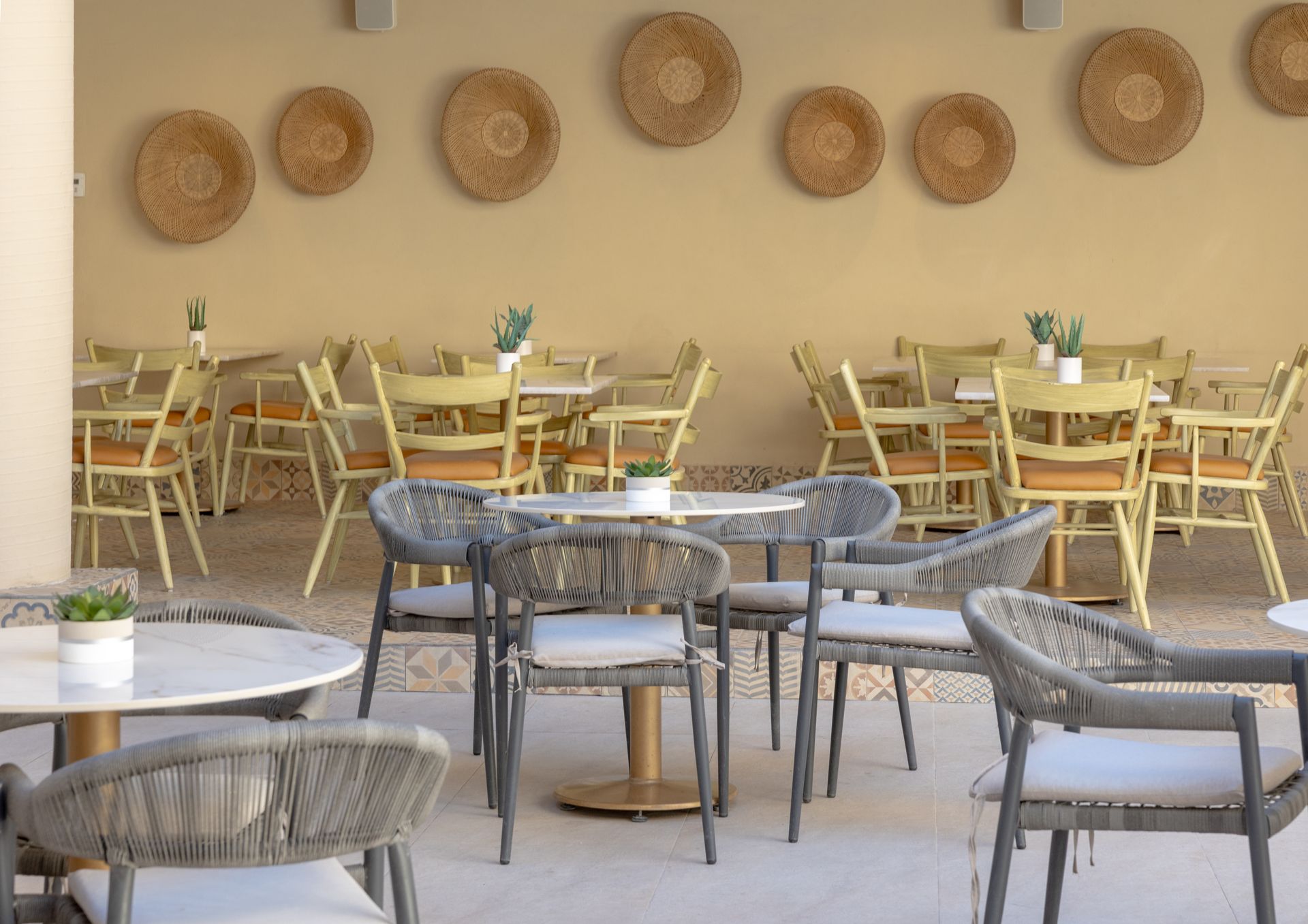 Cafetería al aire libre con mesas redondas y sillas tejidas; sillas amarillas y decoración de pared tejida en el fondo.