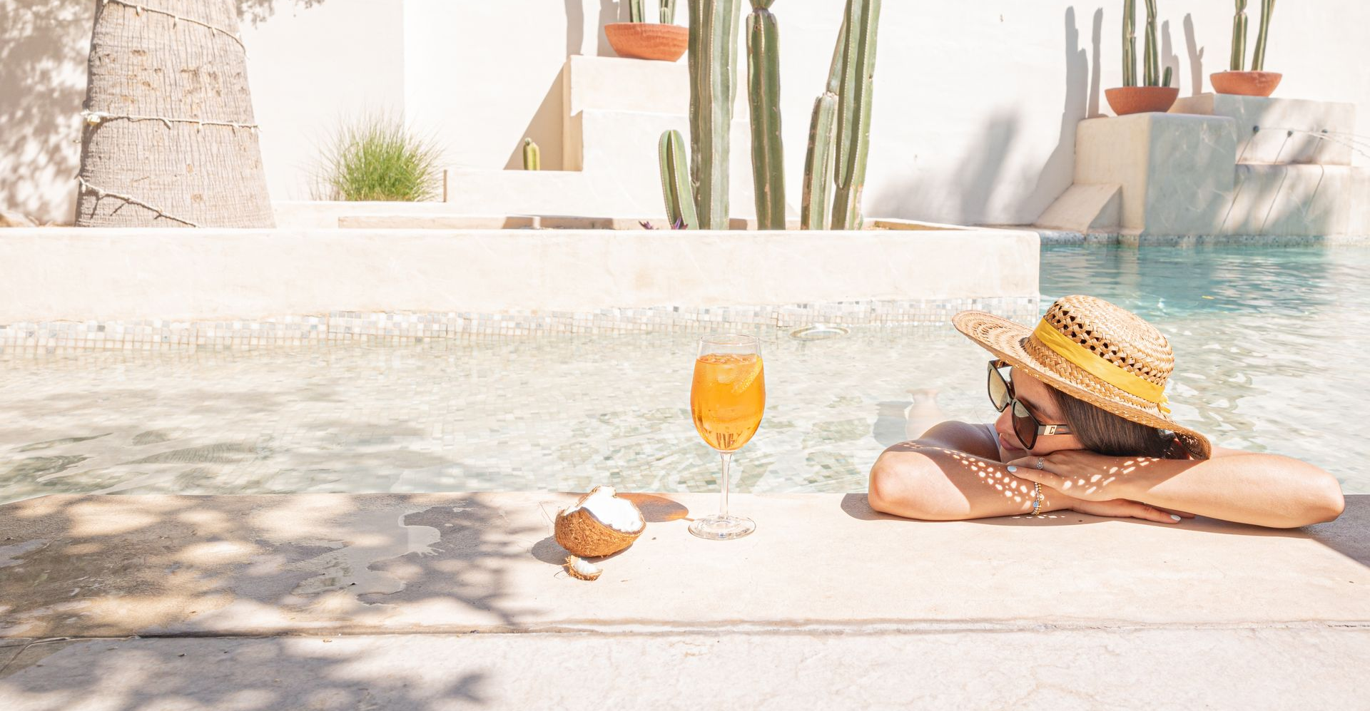 Persona con sombrero de paja junto a una piscina, disfrutando de una bebida y del clima soleado.