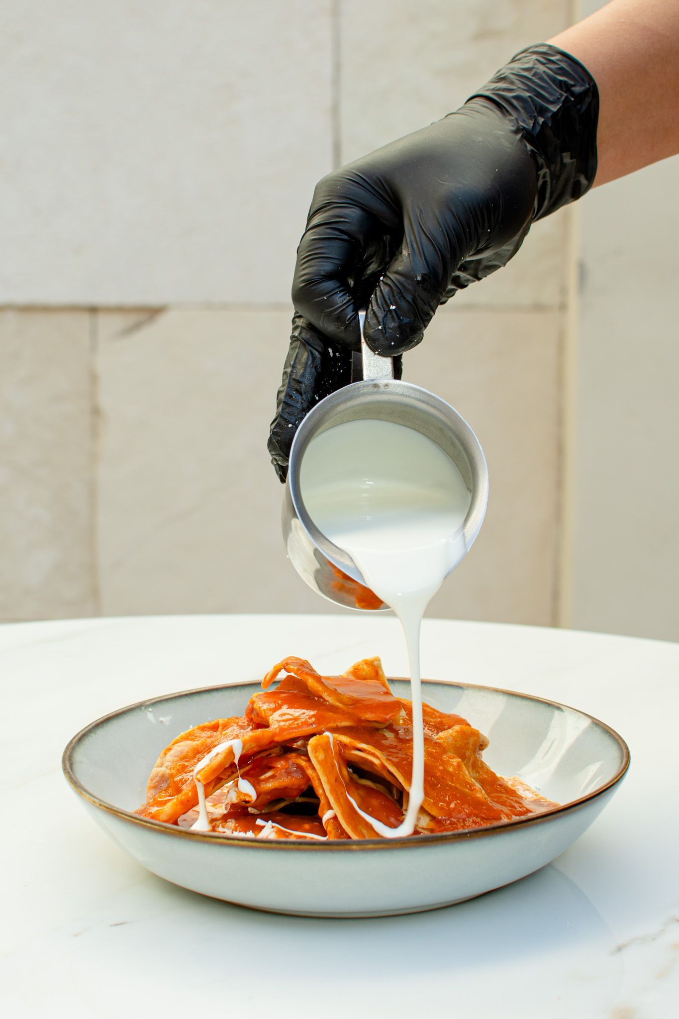 Una persona con guante negro vierte un líquido blanco sobre comida naranja en un recipiente.
