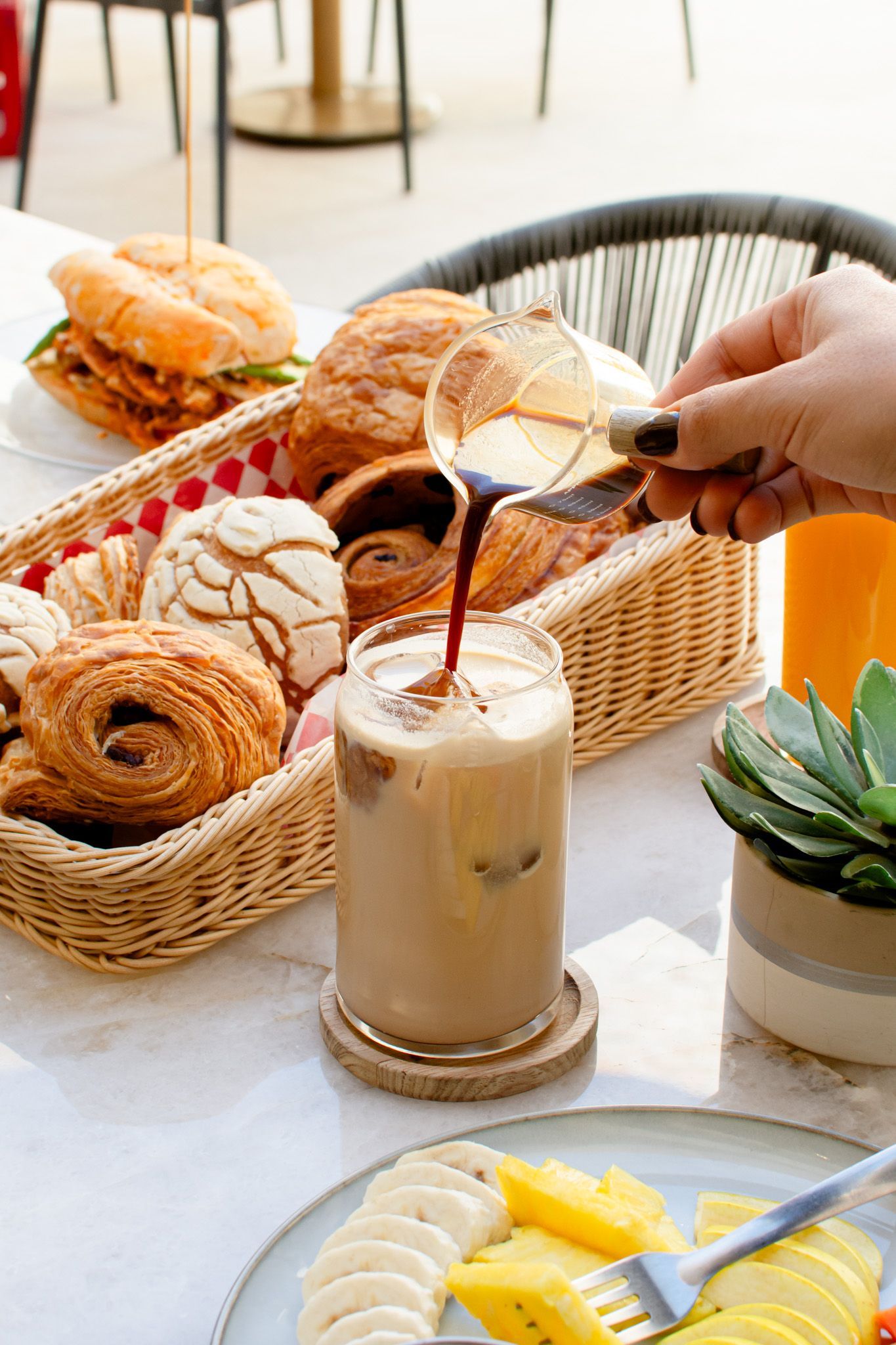 Café servido en un vaso con hielo. Cesta de pasteles, plato de fruta y zumo de naranja sobre la mesa.