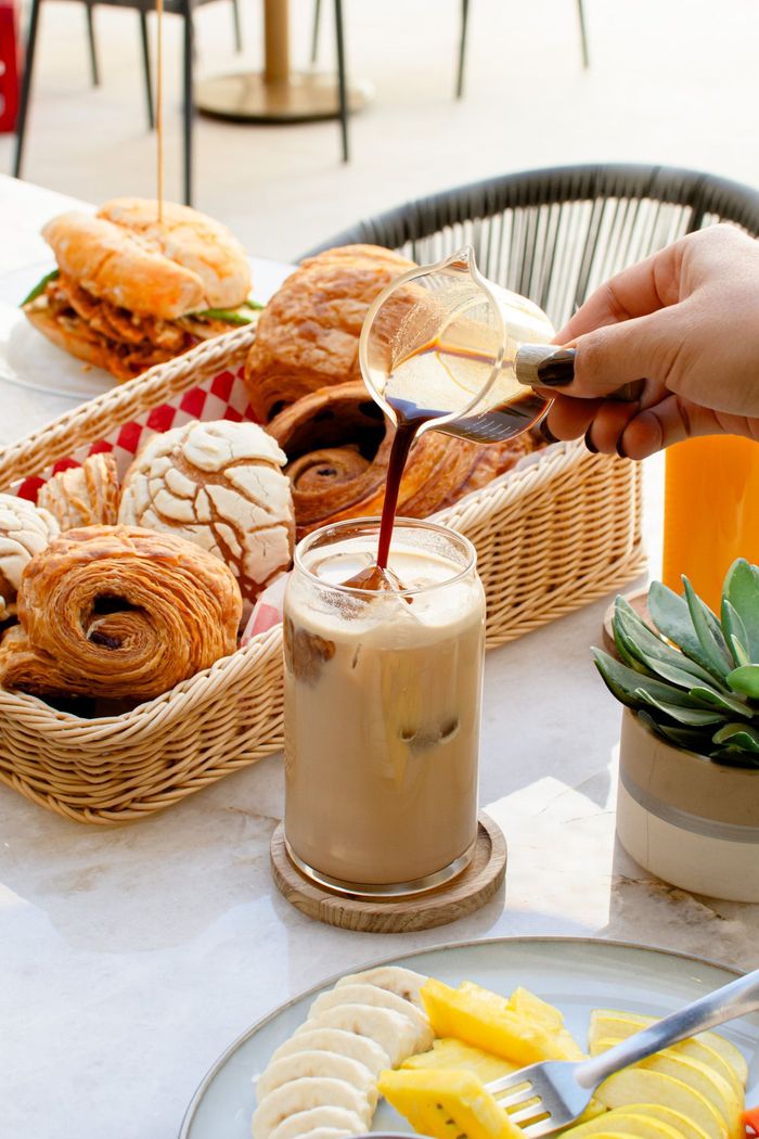 Café servido en un vaso con hielo. Cesta de pasteles, plato de fruta y zumo de naranja sobre la mesa.