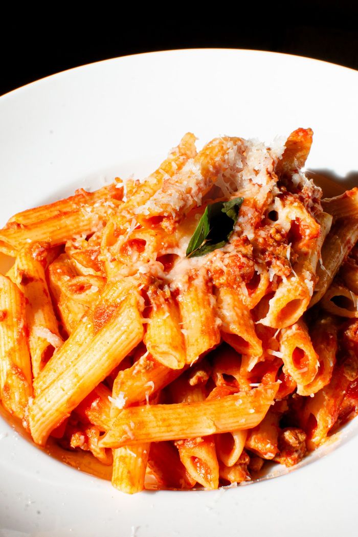 Pasta penne en salsa roja, cubierta con queso rallado y una ramita de albahaca, en un plato blanco.