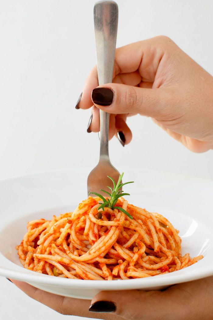 Mano sosteniendo un tenedor mientras hace girar espaguetis con salsa roja, adornados con hierbas, en un plato blanco.