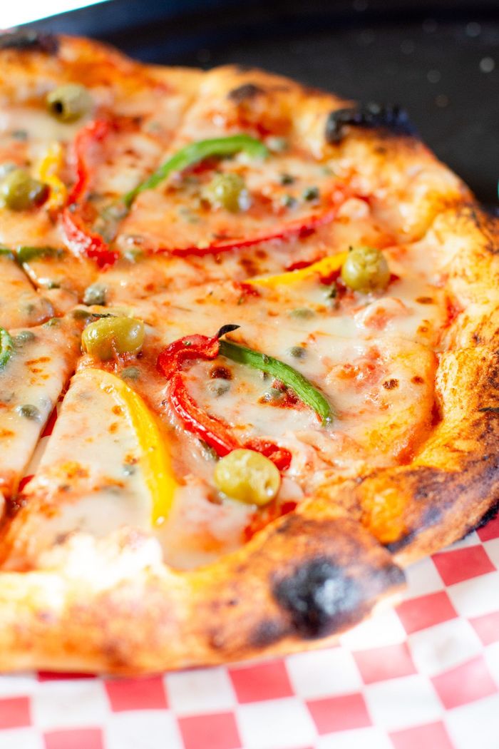 Pizza con pimientos rojos, amarillos y verdes, aceitunas y queso sobre un papel a cuadros rojo y blanco.