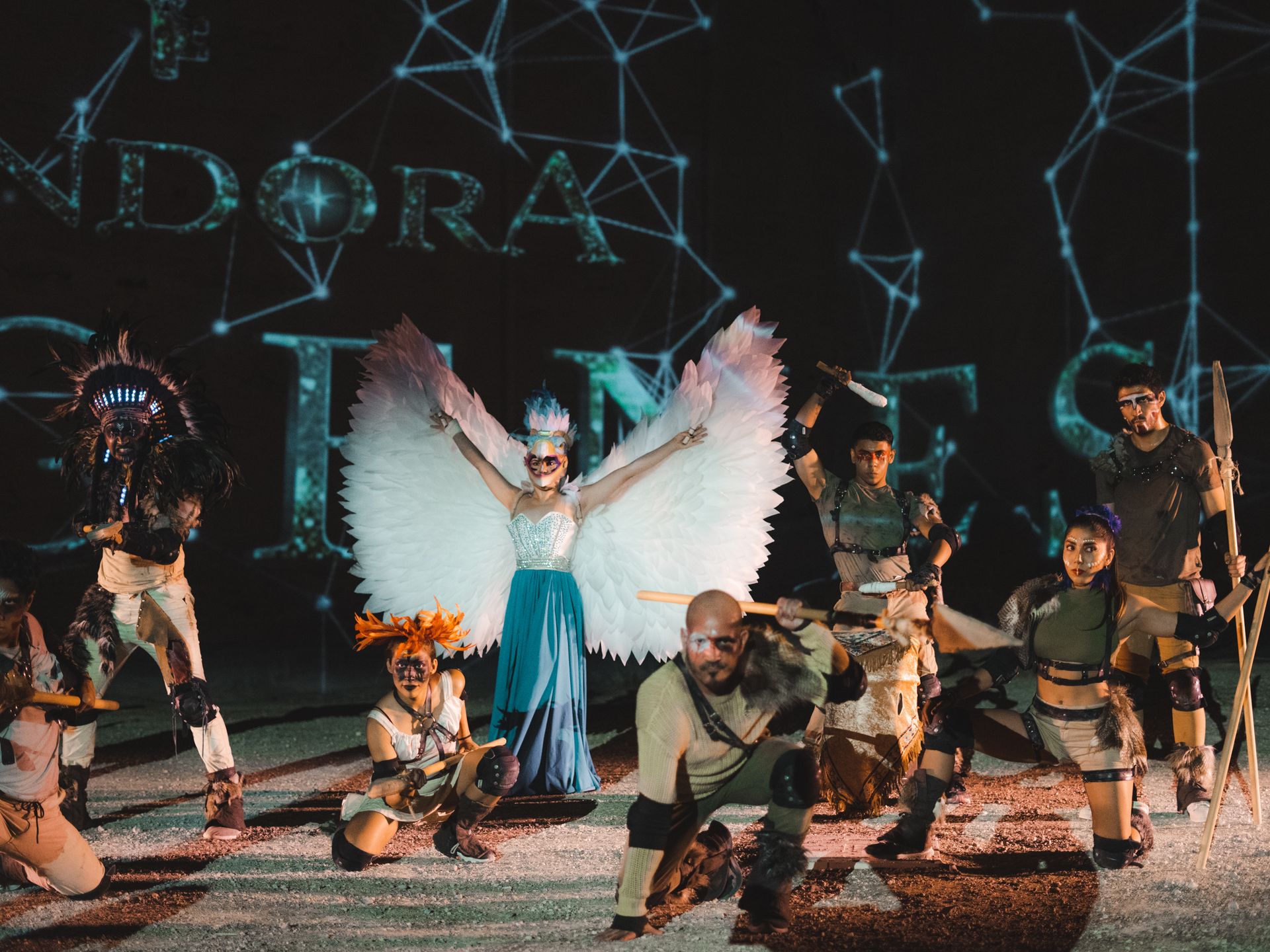 Grupo de artistas disfrazados posando en un escenario con un fondo proyectado, con una figura central con grandes alas blancas.