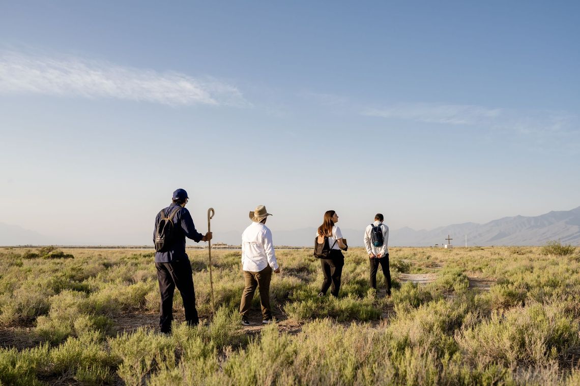 Cuatro personas están de pie en un campo de arbustos verdes bajo un cielo azul, con montañas en la distancia.