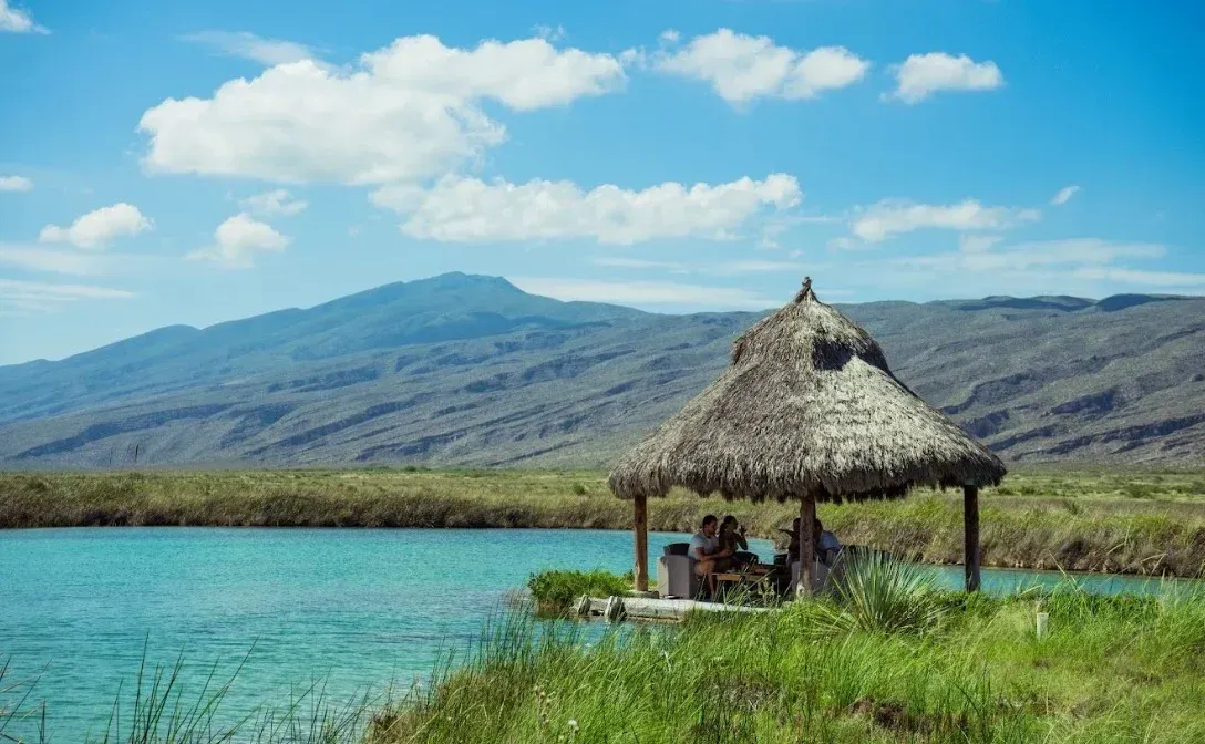 Imagen de un río  turquesa con fondo de unas montañas y al frente una palapa en un día soleado 