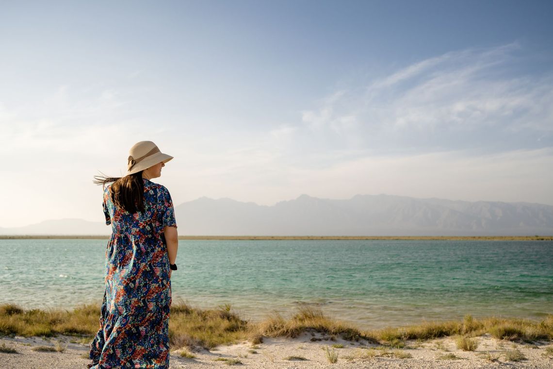Mujer con vestido floral y sombrero mira el agua turquesa y las montañas bajo un cielo azul.