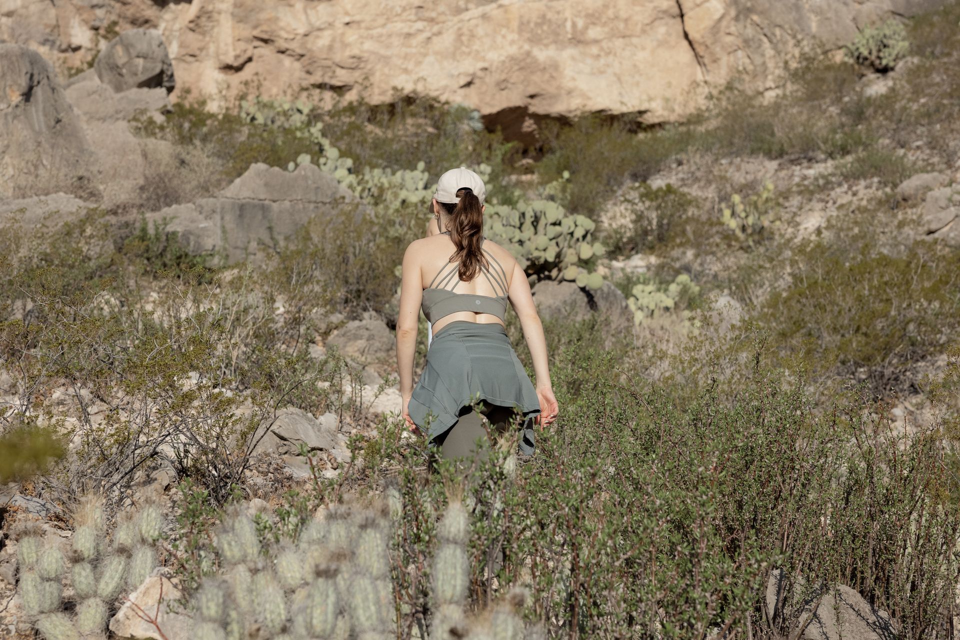 Mujer caminando cuesta arriba en un paisaje desértico, vestida con sombrero, sujetador deportivo y chaqueta.