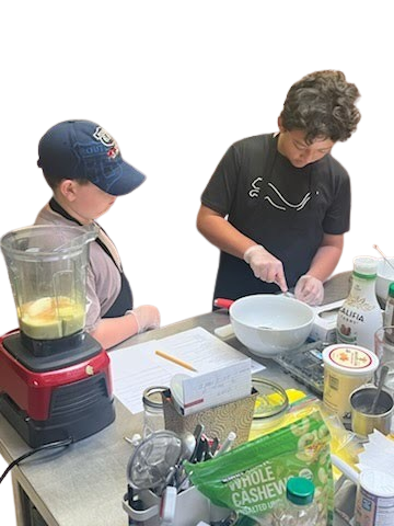 Two students wearing gloves prepare ingredients in a kitchen with a blender, bowl, and snacks on a counter.