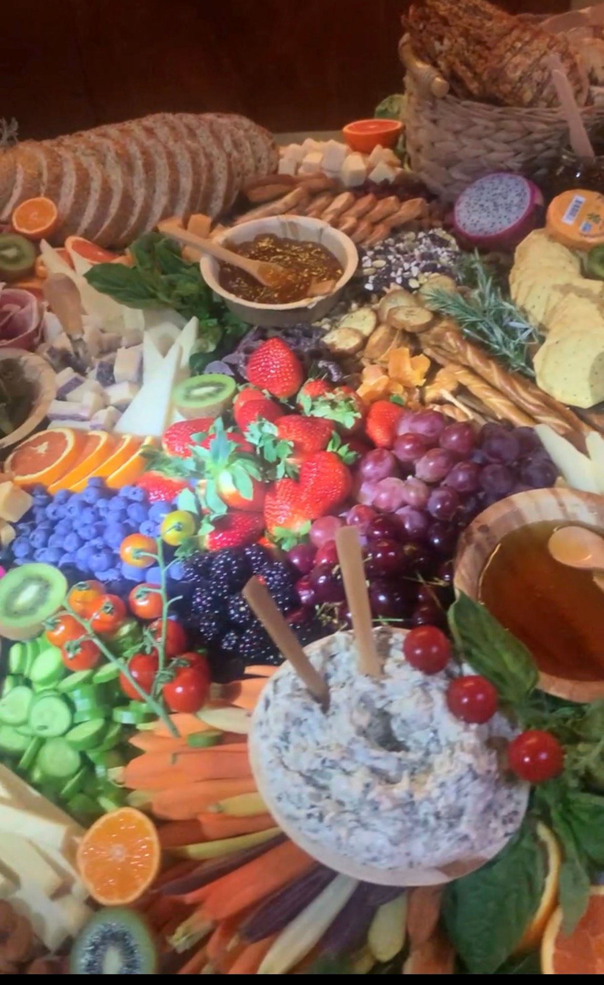A colorful grazing board filled with assorted cheeses, bread slices, fresh fruit, vegetables, nuts, and dipping sauces.