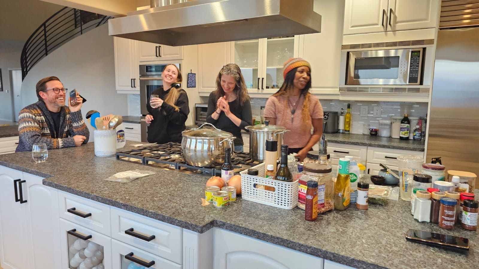 Four people laugh in a bright, modern kitchen while standing behind a large granite island with cooking ingredients.