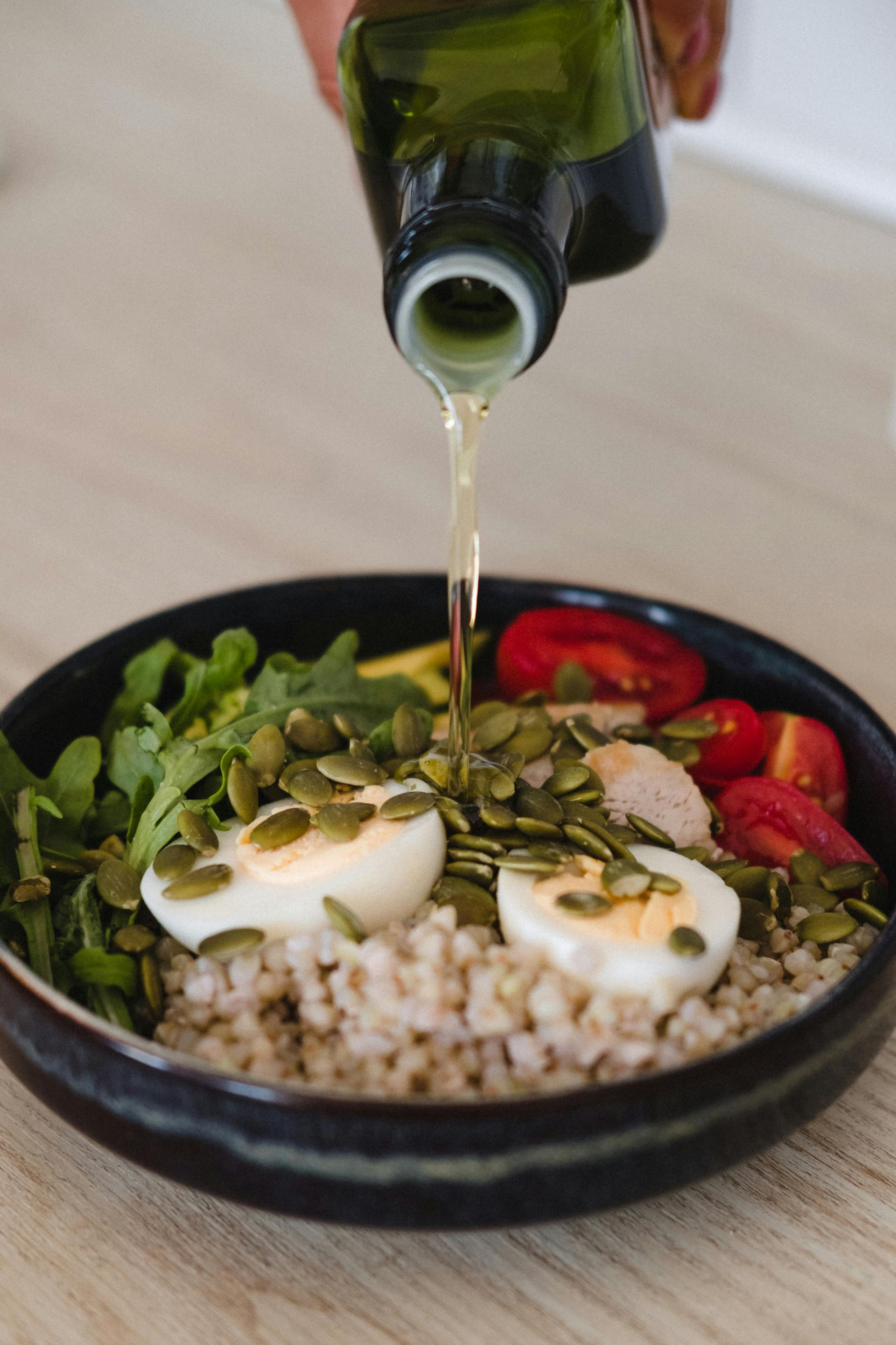 Oil is poured from a glass bottle onto a nutritious bowl containing hard-boiled eggs, seeds, greens, and cherry tomatoes.