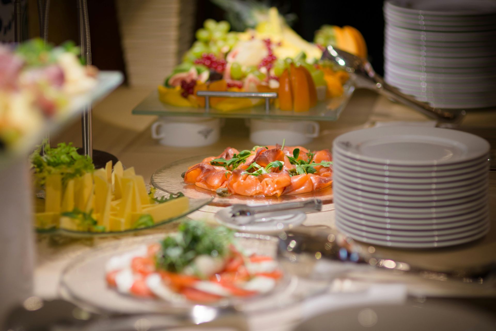 A buffet display featuring platters of sliced fruit, salmon, and caprese salad, with stacks of white plates nearby.