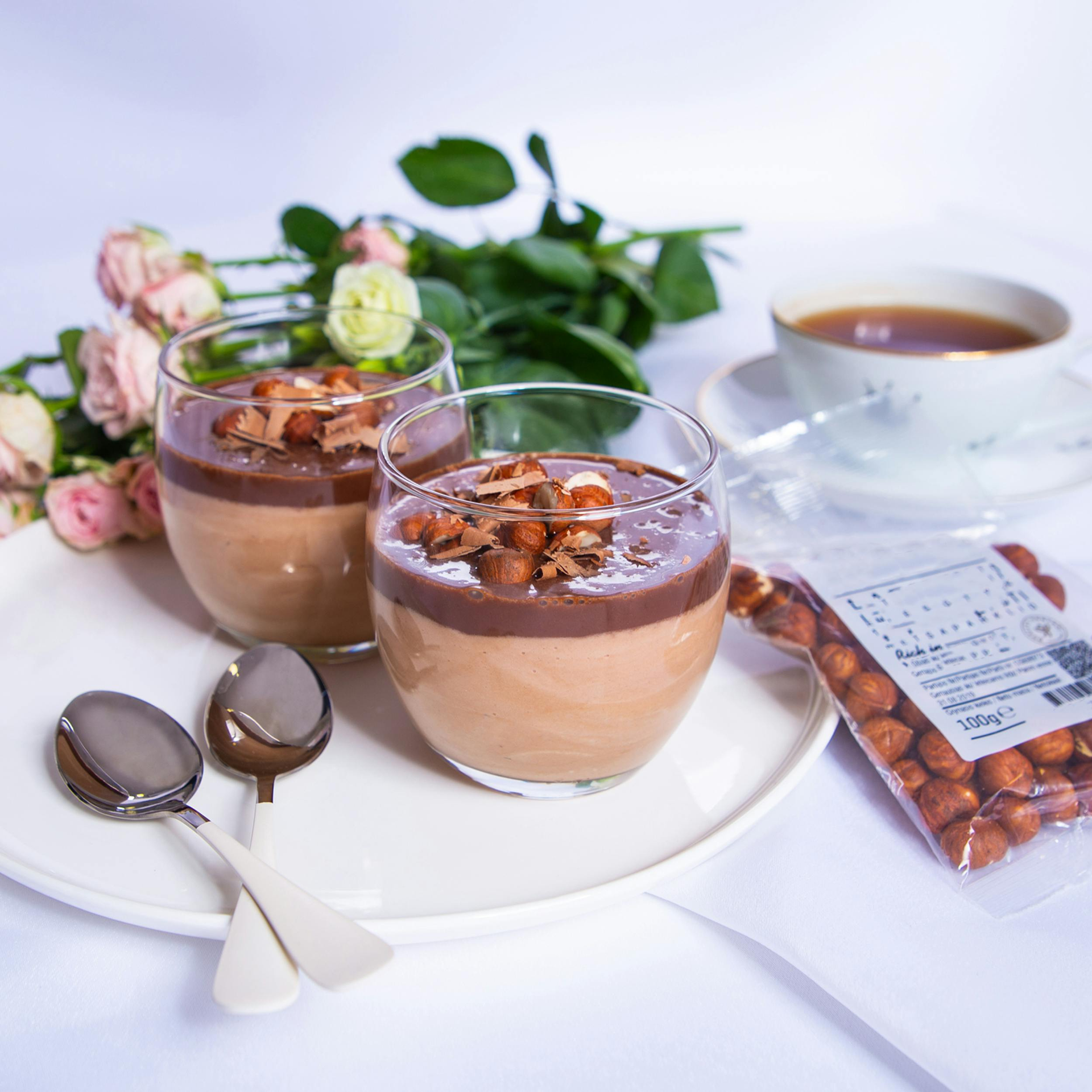 Two glasses of chocolate hazelnut mousse topped with nuts and shavings, served with spoons, flowers, and a cup of tea.