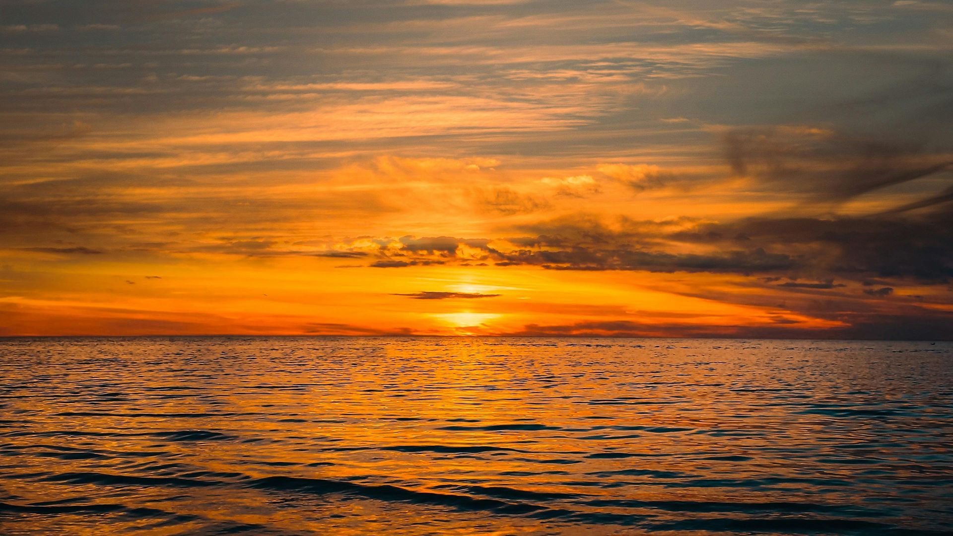 A vibrant orange sunset glows over the horizon, casting shimmering reflections across the textured surface of the water.