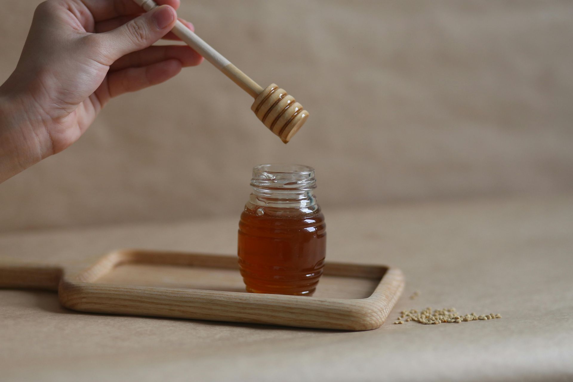 A hand holds a wooden honey dipper over a small glass jar of golden honey resting on a wooden tray against a beige background.