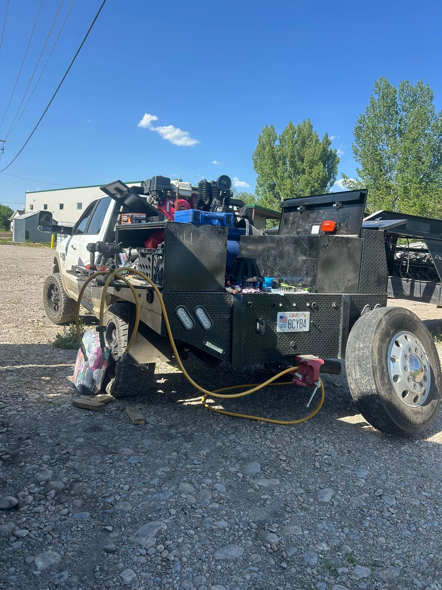 A black utility truck with equipment and tools parked on gravel under a blue sky. A white building is visible in the background.