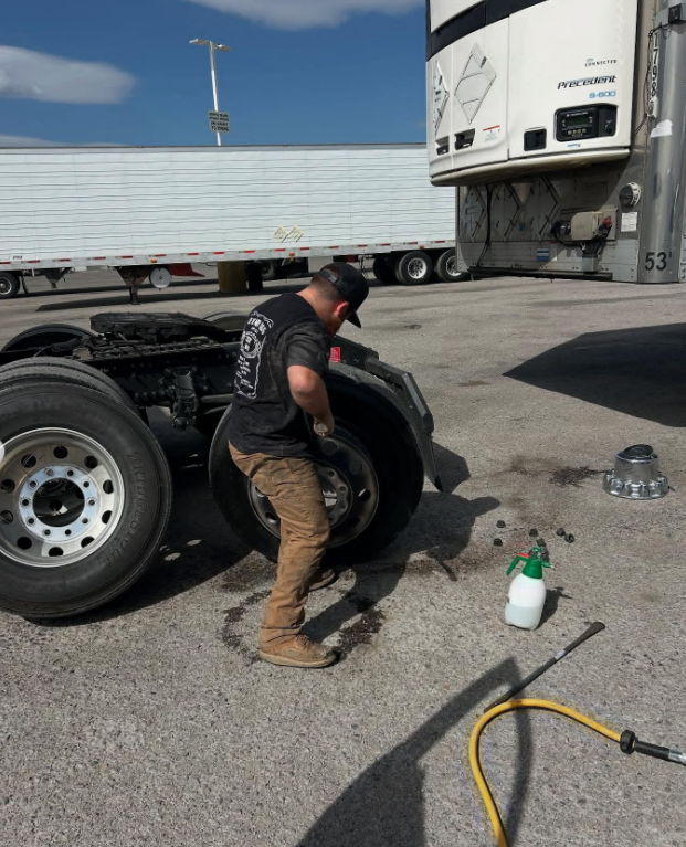 A person in work clothes changes a tire on a semi-truck in a paved lot on a sunny day.