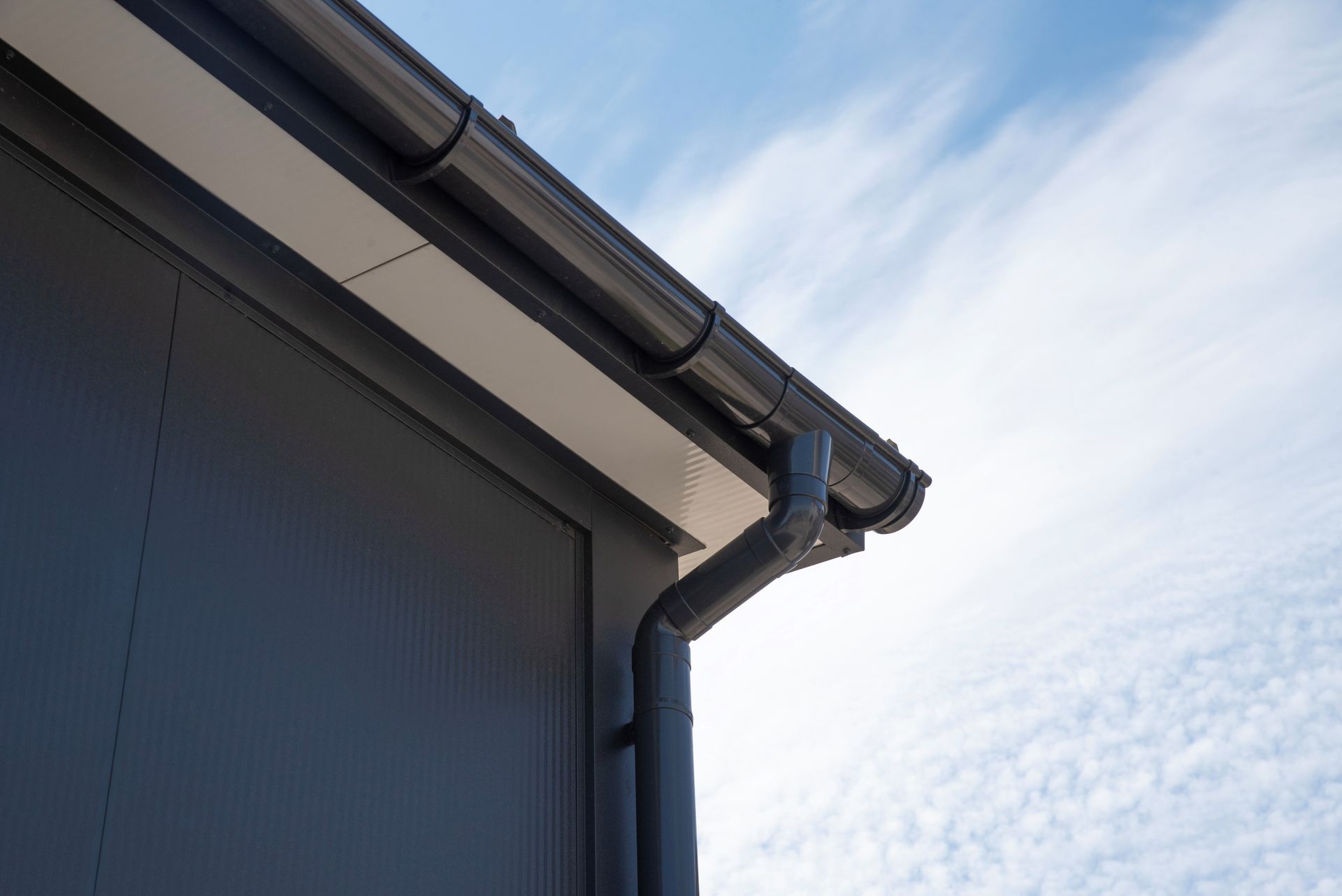 Close-up of a gray building roof and gutter against a blue sky with wispy clouds