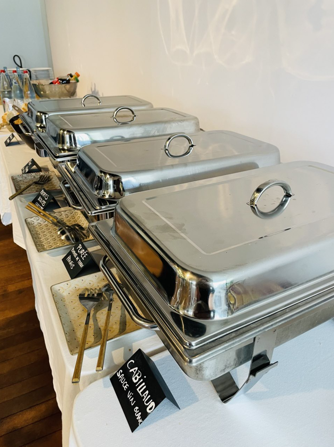 Buffet avec chauffe-plats couverts en acier inoxydable, étiquettes des aliments sur une nappe blanche.