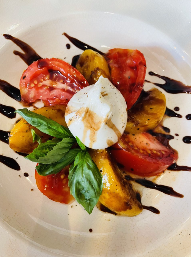 Salade Caprese : tomates rouges et jaunes tranchées, fromage burrata, basilic, glaçage balsamique, sur assiette blanche.