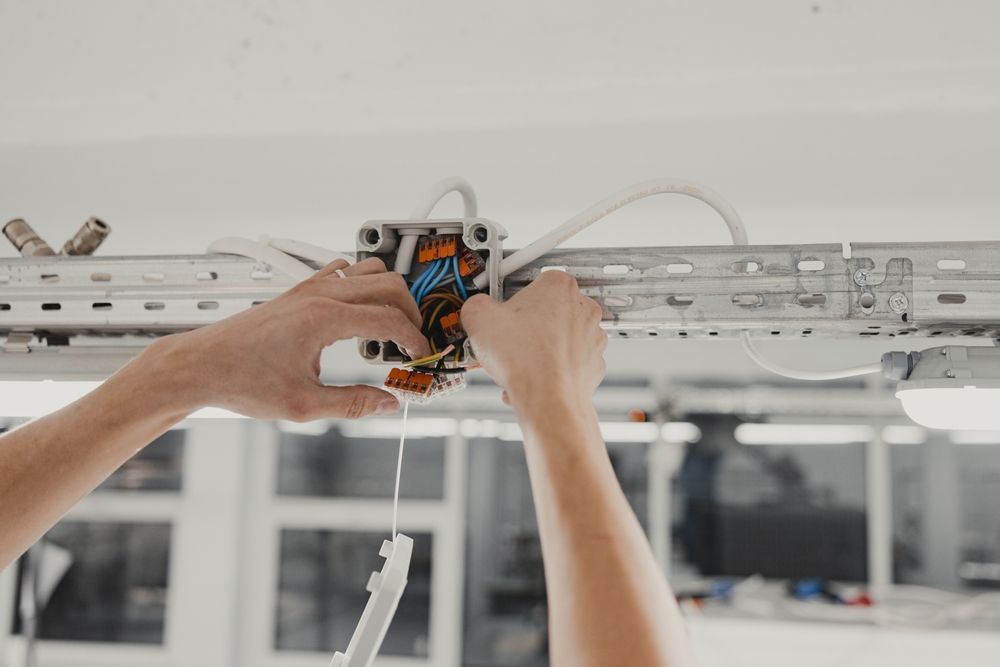 Hands wiring a ceiling-mounted electrical box with exposed cables and white conduit in a bright room