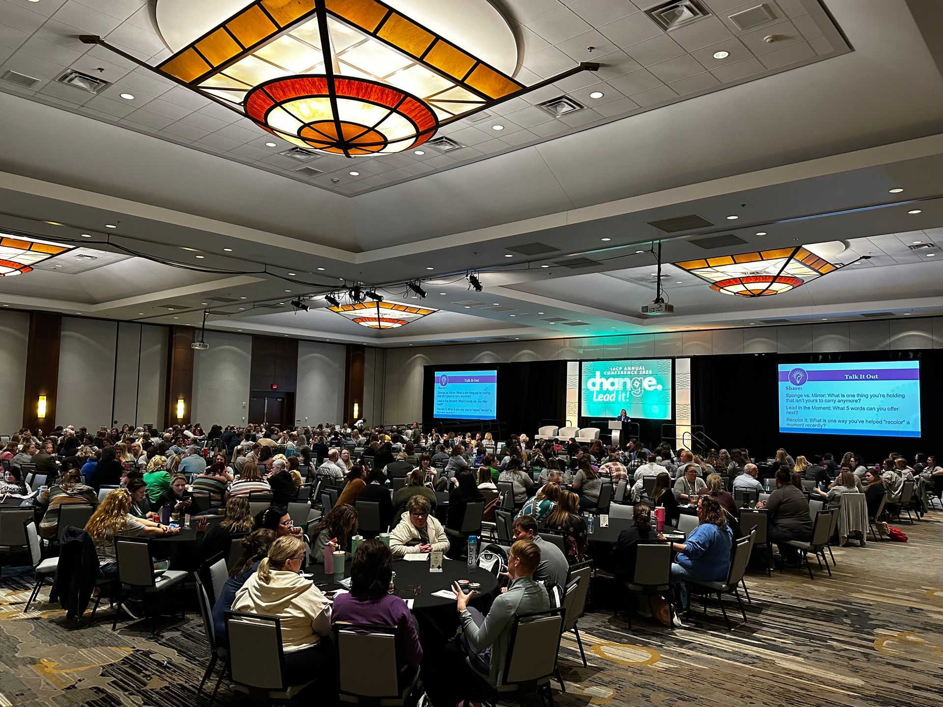 Conference ballroom packed with seated attendees facing a stage with large presentation screens