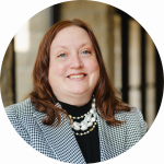 Smiling woman with auburn hair in a black-and-white checkered blazer and pearl necklace against a blurred background