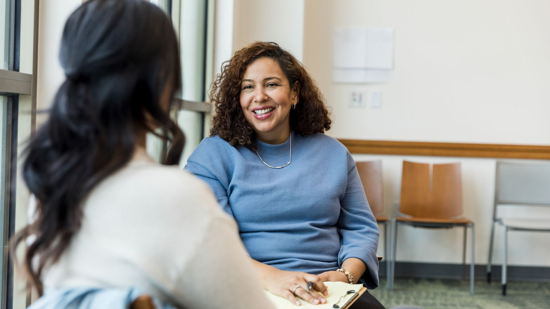 Two women talking in a bright office, one smiling while seated in a blue sweater.