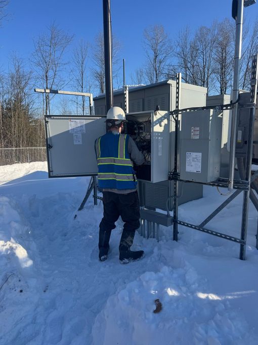 A worker in a high-visibility vest and hard hat services an open electrical box in a snowy, outdoor industrial setting.